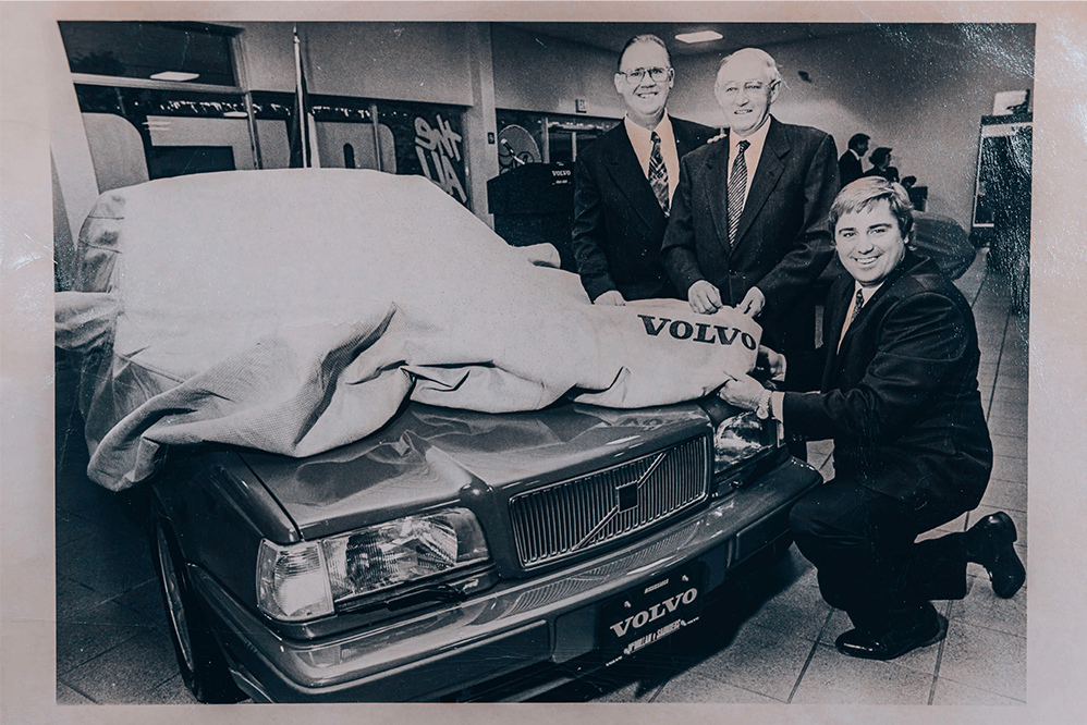 A black-and-white photo of three men in suits unveiling a new Volvo sedan, which is partially covered by a canvas sheet bearing the Volvo logo.