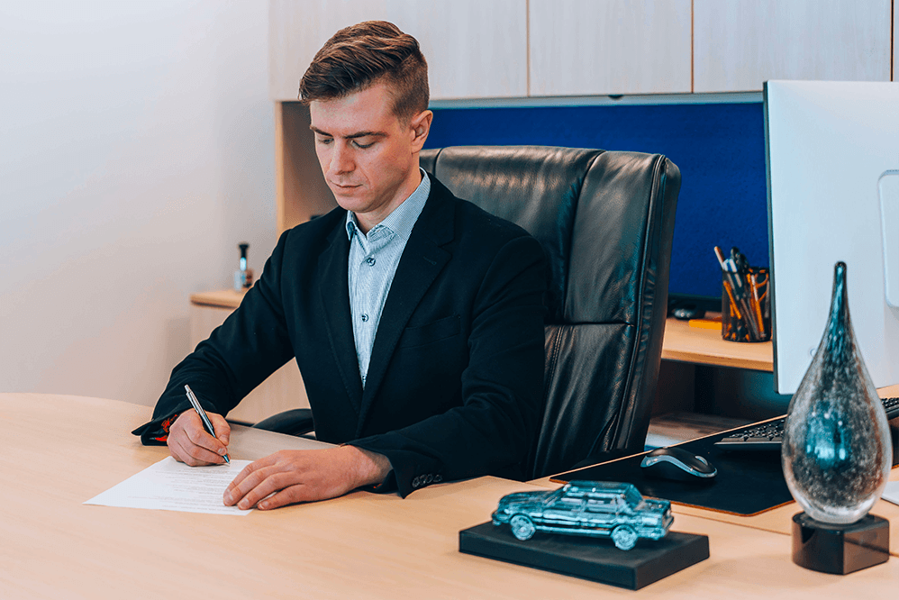 A man in a black blazer and light blue dress shirt sitting at a wooden desk and signing a document.