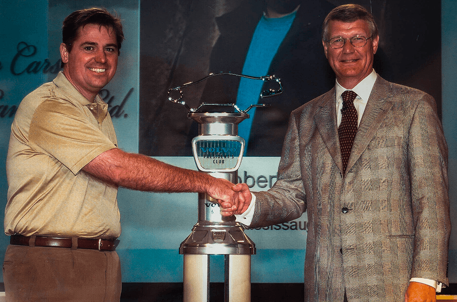 An indoor photograph of two smiling men shaking hands across a large metal trophy. The trophy features a blue section inscribed with "VOLVO PRESIDENT'S CLUB" and is topped by a car silhouette.