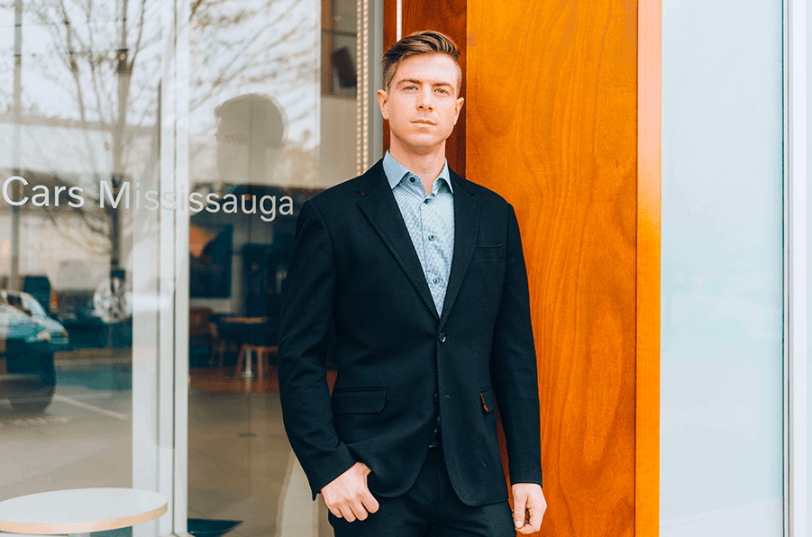A man in a black suit jacket and blue patterned shirt standing outside a dealership building next to a glass window with "Cars Mississauga" text.