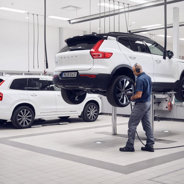 A technician wearing earmuffs works on the wheel of a white Volvo SUV on a lift in a bright, clean service garage.