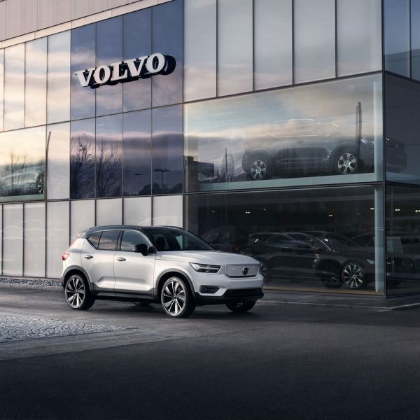A white Volvo XC40 parked in front of a modern, multi-story glass Volvo dealership building at dusk.