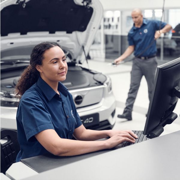 A female Volvo service employee in a blue uniform types at a computer, while a male technician works on a white SUV with its hood open in the background.