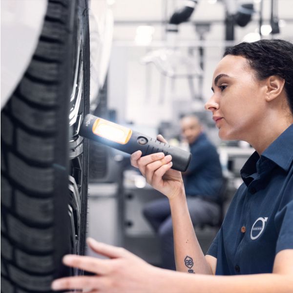 A female Volvo service technician closely inspects a car's tire tread using a bright handheld work light.