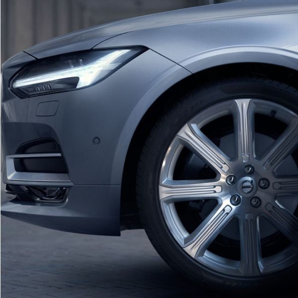 A close-up shot of the front headlight and multi-spoke alloy wheel of a silver Volvo sedan.