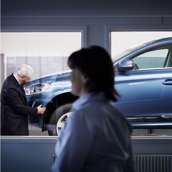 A man in a suit inspects the front wheel of a blue Volvo SUV on a lift in a clean service bay, viewed from behind a window.