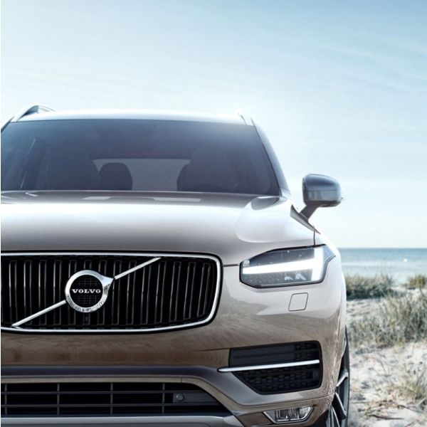 A close-up of the front grille and "Thor's Hammer" headlights of a modern Volvo SUV parked on a sandy beach, with the ocean in the background.