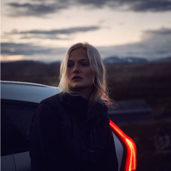 A blonde woman in a dark jacket stands next to a car in a remote, hilly landscape at dusk, illuminated slightly by the car's red taillight.