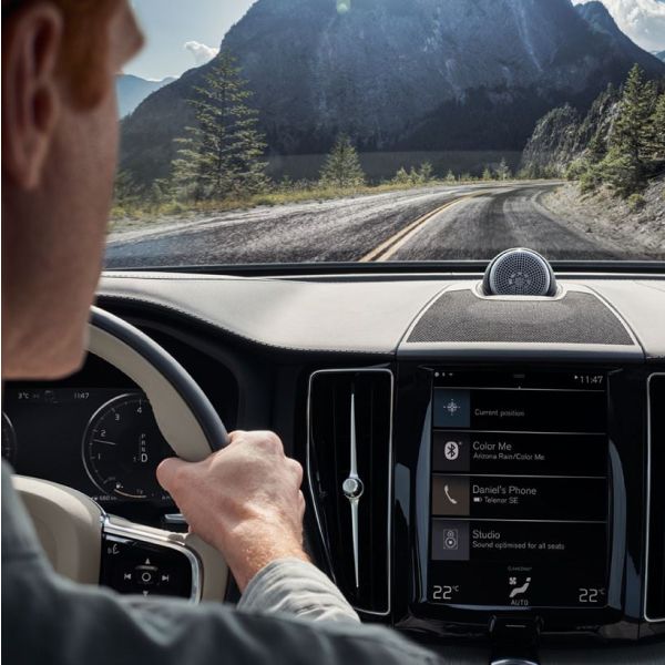 An over-the-shoulder view from inside a Volvo, showing the driver's hands on the steering wheel, the center touchscreen, and a mountain road ahead.