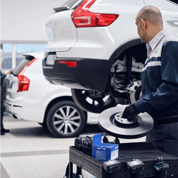 A Volvo service technician in uniform holds a new brake disc in a garage. A cart with a 'Volvo Genuine Parts' box is nearby, and a white Volvo SUV is on a lift.