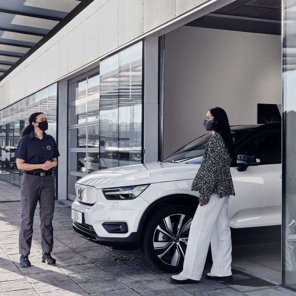 A female Volvo service employee and a female customer, both wearing face masks, stand and talk next to a white Volvo SUV outside a service center.
