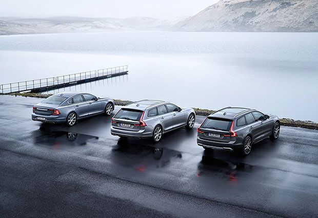 Three silver Volvo models (S90 sedan, V90 wagon, and V90 Cross Country) parked in a row on wet pavement next to a large, tranquil lake and hills.