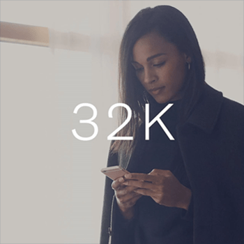 A woman in a dark coat stands indoors near a bright window, looking down at her smartphone, with the text "32K" overlaid.