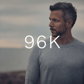 A profile view of a man in a grey sweater standing outdoors with a blurred coastline in the background, with "96K" text overlaid.