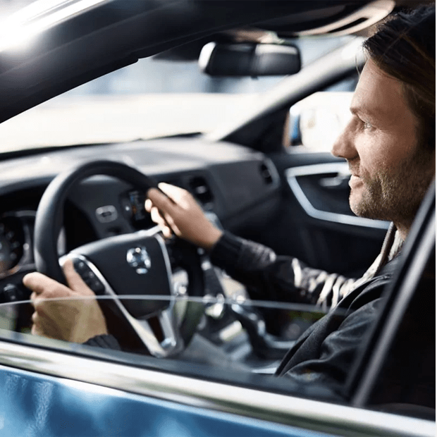 A smiling man seen through the driver's side window, with his hands on the steering wheel, driving a modern car.