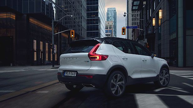 A rear-quarter view of a white Volvo XC40 SUV with a black roof, driving on a city street surrounded by tall buildings.