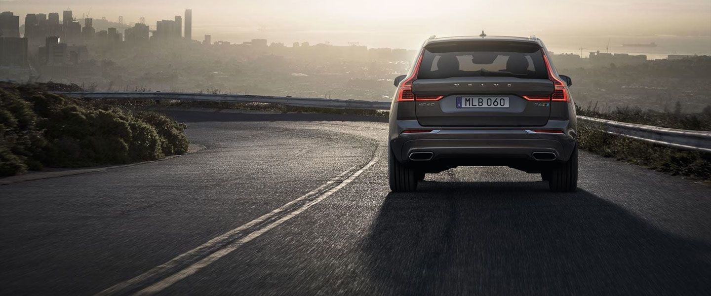 A rear view of a dark grey Volvo XC60 SUV driving on a winding road, overlooking a hazy city skyline in the distance during sunrise or sunset.