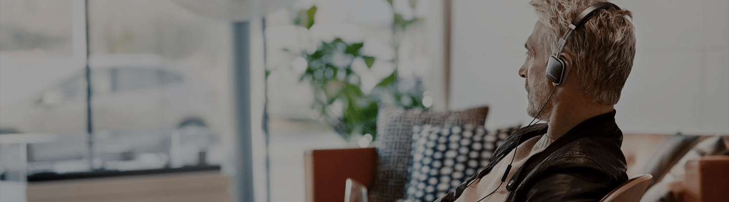 A man with salt-and-pepper hair sits in a modern, light-filled lounge, looking thoughtfully to the side while listening to on-ear headphones.