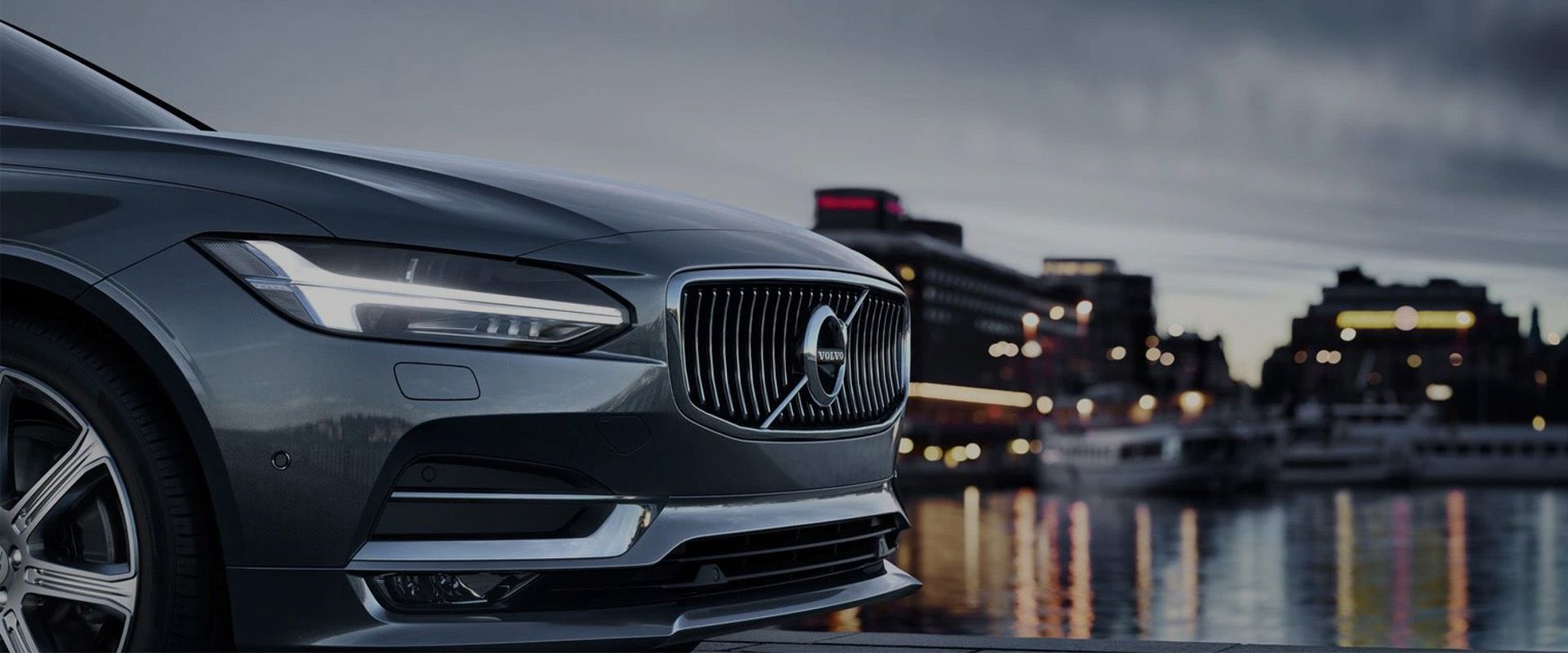 The front quarter of a dark grey Volvo sedan parked next to a body of water at dusk, with a blurred city skyline and lights in the background.