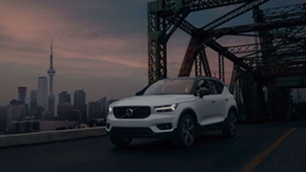 A white Volvo XC40 SUV driving across a metal truss bridge at dusk, with the Toronto skyline, including the CN Tower, visible in the background.