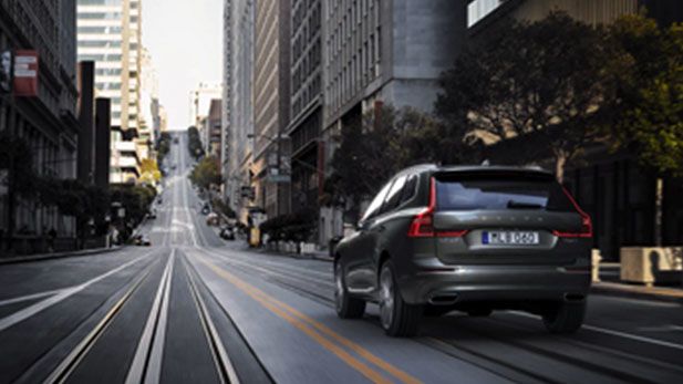 A rear view of a dark grey Volvo XC60 SUV driving down a steep, multi-lane city street with cable car tracks.