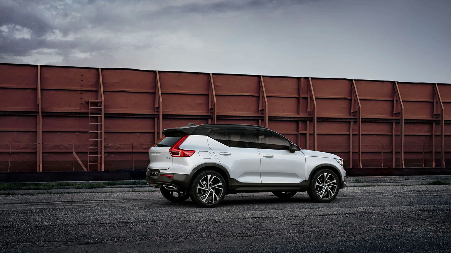 A side-rear view of a white Volvo XC40 with a black roof, parked on dark pavement in an industrial area in front of a long, red-brown train.
