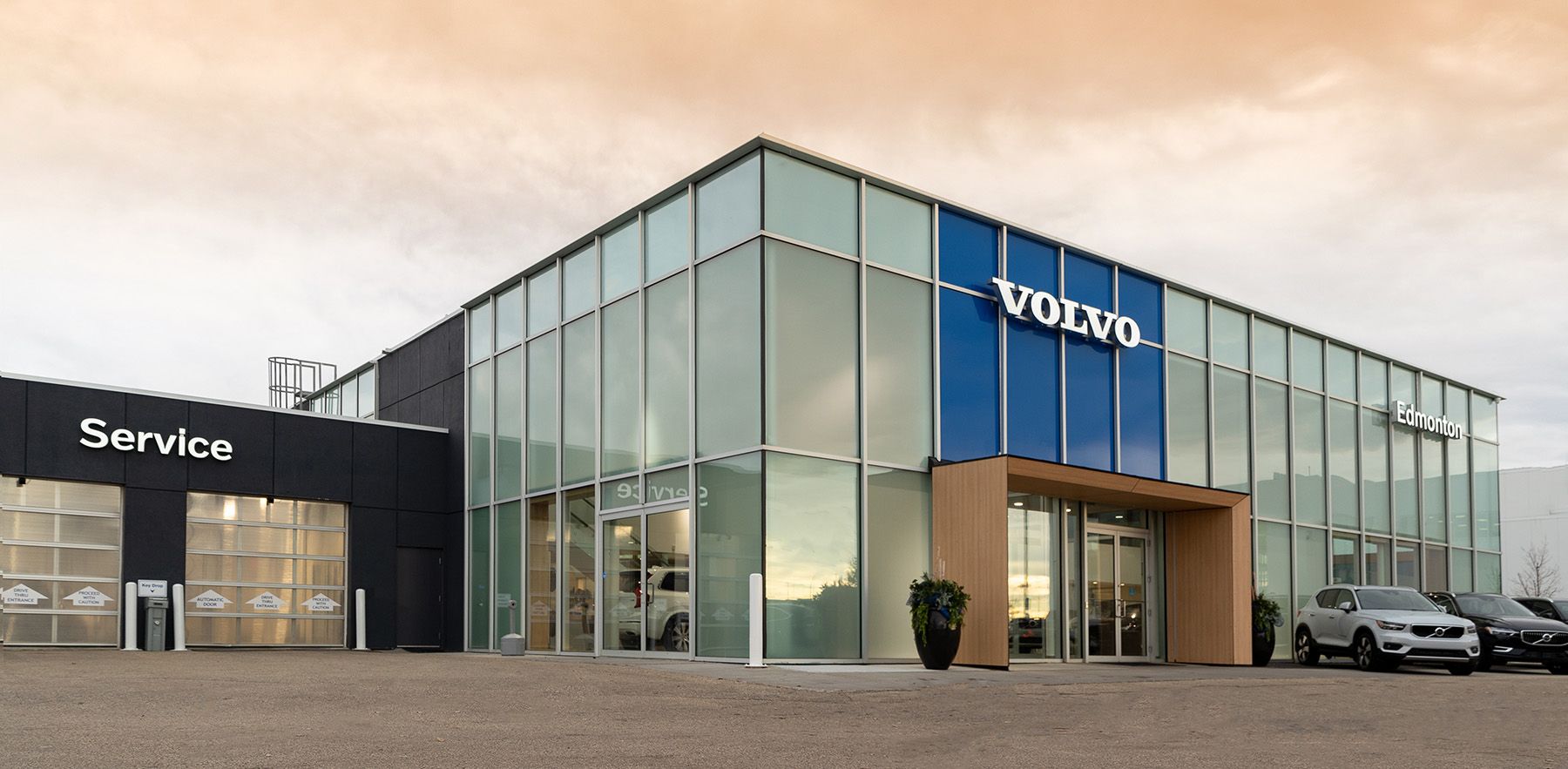 Exterior view of the Volvo of Edmonton dealership, featuring a modern glass facade with a large blue Volvo logo, a designated Service center entrance on the left, and new vehicles on display in front.
