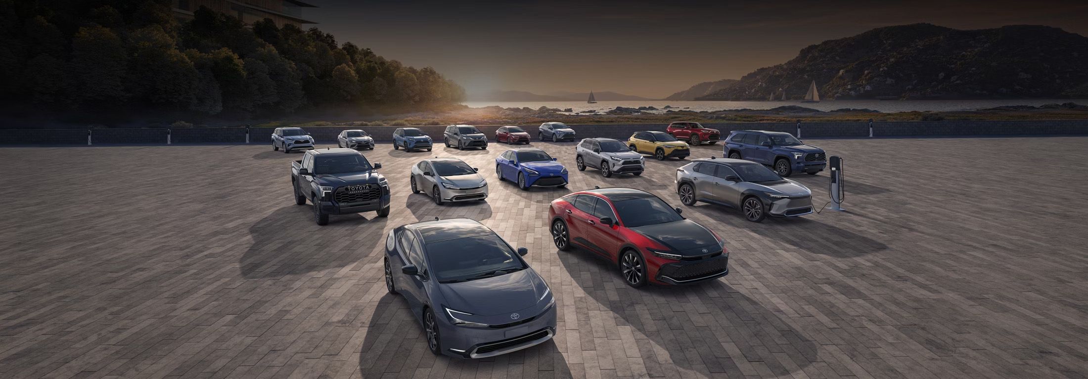 A diverse lineup of new Toyota vehicles, including SUVs, sedans, and trucks, parked on a large paved area near a body of water with mountains and trees in the background under a dramatic sky. Many vehicles appear to be electrified models, some with charging stations nearby.