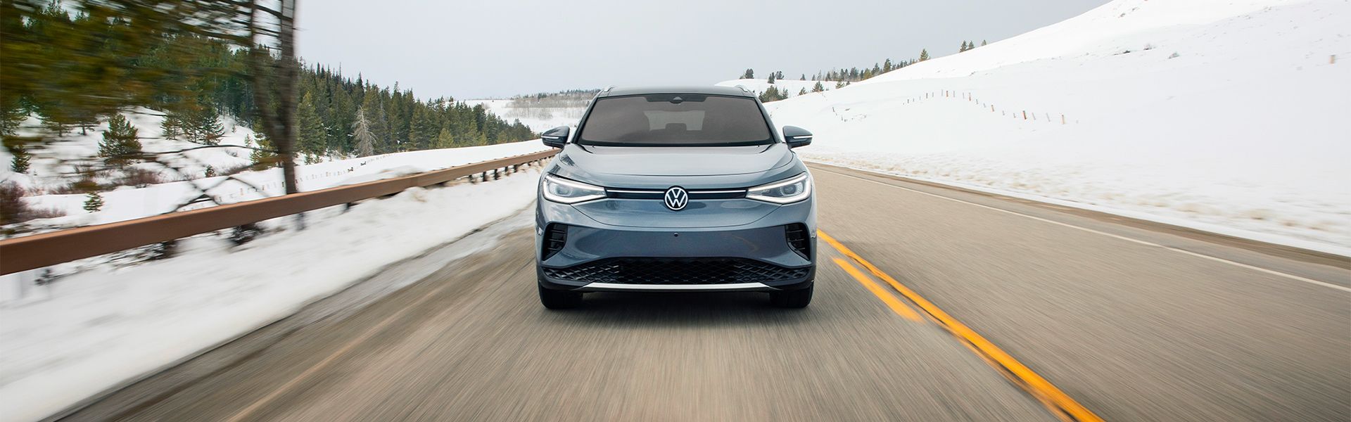 A front-facing view of a slate blue Volkswagen ID.4 driving down a paved mountain road flanked by snow-covered hills and pine trees. The motion blur on the road and trees creates a sense of speed and adventure in a winter landscape.