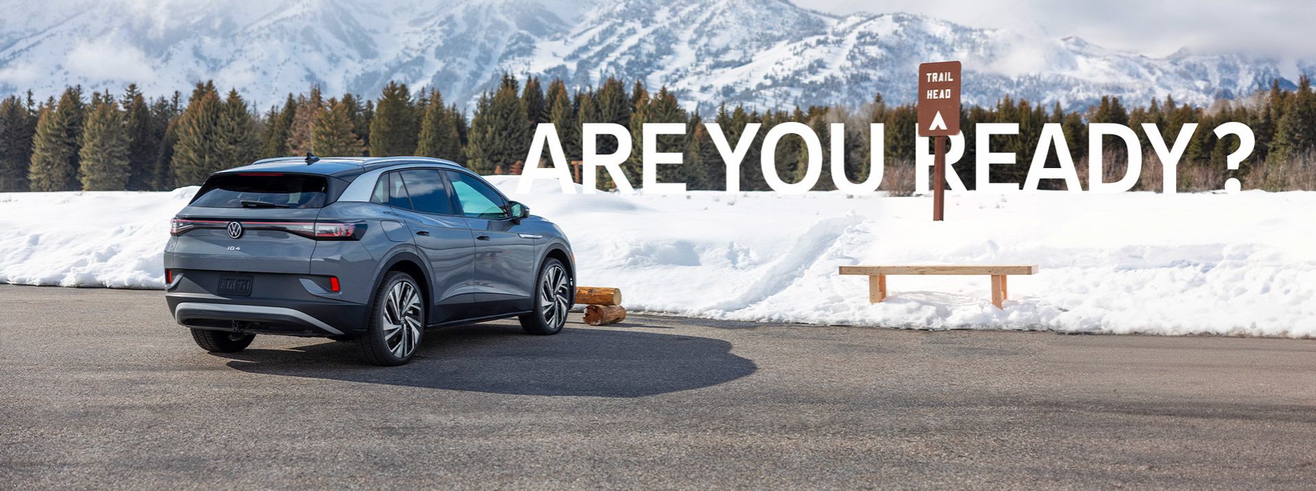 A three-quarter rear view of a slate blue Volkswagen ID.4 parked in a snowy mountain lot next to a wooden bench and a "Trail Head" sign. Large, bold white text overlaid on the snowy background asks, "ARE YOU READY?" with majestic, snow-capped peaks in the distance.