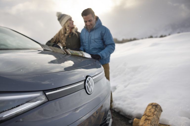 A close-up of the front hood of a grey Volkswagen, focusing on the silver VW logo. In the blurred background, a man and a woman in winter parkas and hats are leaning over the car, looking at a map together in a snowy outdoor setting.