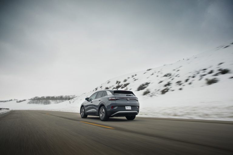 A rear three-quarter view of a grey Volkswagen ID.4 traveling along a winding asphalt road. The vehicle is surrounded by a vast, rolling landscape of snow-covered hills under a soft, overcast winter sky.