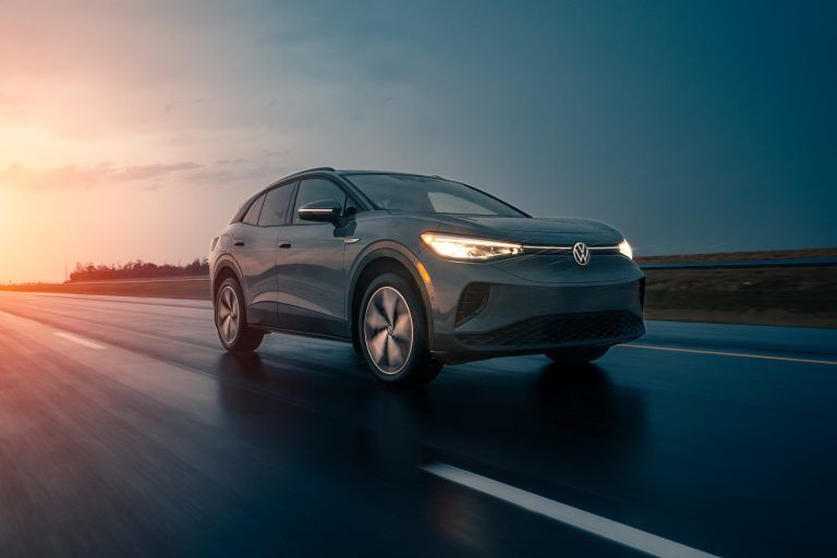 A sleek, grey Volkswagen ID.4 driving along a highway during a dramatic sunset. The warm orange light of the setting sun glows on the horizon, reflecting off the damp road surface and highlighting the car's modern LED headlights and illuminated front logo.
