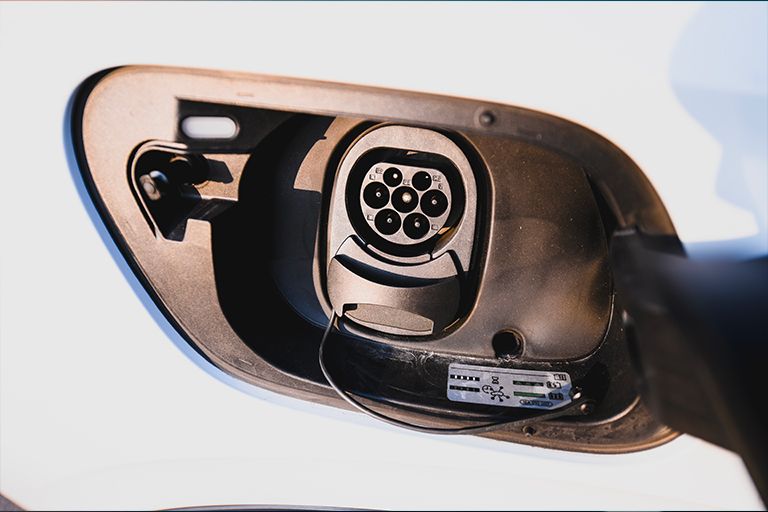A close-up, high-angle shot of the open charging port on a white Volkswagen vehicle. The circular port features a seven-pin Type 2 connector design housed within a dark, protective plastic compartment.